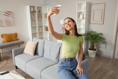 Genç bir kadın oturma odasındaki gri kanepede selfie çekiyor.