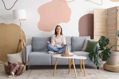 Genç bir kadın oturma odasındaki gri kanepede meditasyon yapıyor.