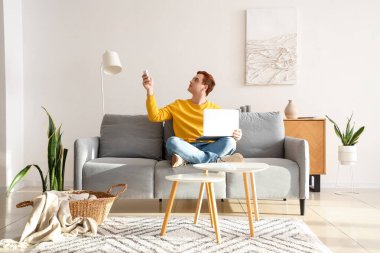 Dizüstü bilgisayarı olan genç adam oturma odasındaki kanepede klimayı açıyor.