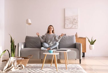 Gözlüklü genç kadın evdeki gri kanepede meditasyon yapıyor.