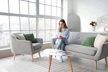 Oturma odasındaki gri kanepede çay içen güzel genç bir kadın.