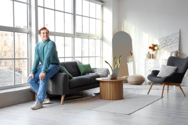 Genç adam oturma odasındaki pencerenin yanındaki yumuşak kanepede oturuyor.