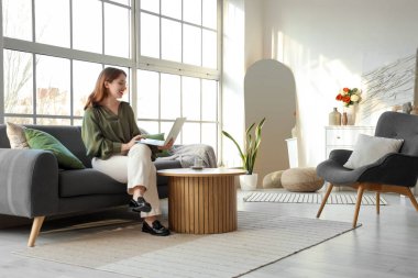 Oturma odasındaki pencerenin yanındaki koltukta dizüstü bilgisayar kullanan genç bir kadın.