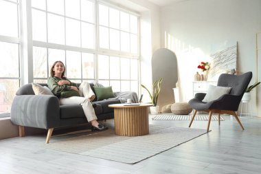 Oturma odasındaki pencerenin yanındaki kanepede uzanan yastıklı genç kadın.