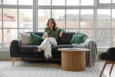 Oturma odasındaki pencerenin yanındaki koltukta cep telefonu kullanan genç bir kadın.