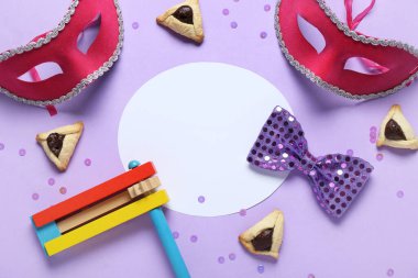 İçinde hamantaschen kurabiyeleri, karnavalın maskeleri ve Purim tatili için mor arka planda çıngırak olan boş bir kart.