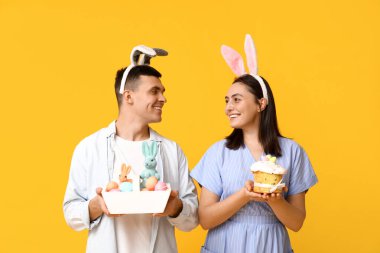 Paskalya tavşanı kulaklı mutlu genç çift, sarı arka planda sepet ve pasta.
