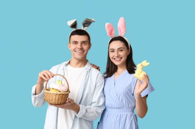 Mavi arka planda Paskalya tavşanı kulaklı, sepetli ve oyuncak tavşanlı mutlu genç çift