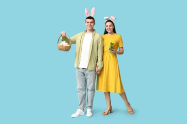 Mavi arka planda Paskalya tavşanı kulaklı, sepetli ve oyuncak tavşanlı mutlu genç çift