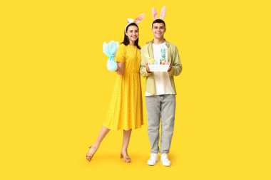 Paskalya tavşanı kulaklı mutlu genç çift, sarı arka planda sepet ve hediye.