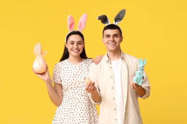 Paskalya tavşanı kulaklı mutlu genç çift, hediye, oyuncak tavşan ve sarı arka planda yumurta.