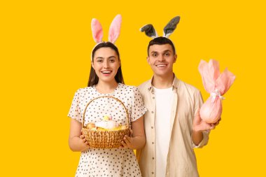 Paskalya tavşanı kulaklı mutlu genç çift, sarı arka planda sepet ve hediye.