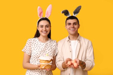 Paskalya tavşanı kulaklı mutlu genç çift, sarı arka planda yumurta ve pasta.