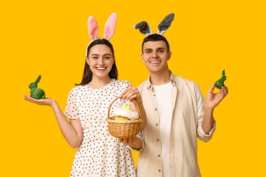 Paskalya tavşanı kulaklı, sepetli ve sarı arka planda oyuncak tavşanları olan mutlu bir çift.
