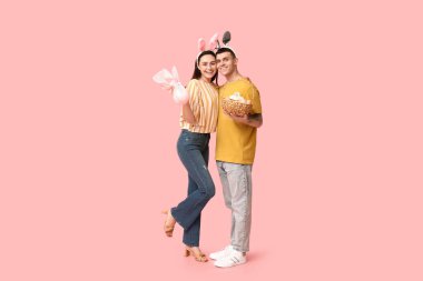 Paskalya sepeti, tavşan kulakları ve pembe arka planda hediyeleri olan mutlu genç çift.