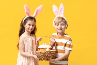 Tavşan kulaklı şirin çocuklar. Paskalya pastası sepeti ve sarı arka planda yumurtalar.