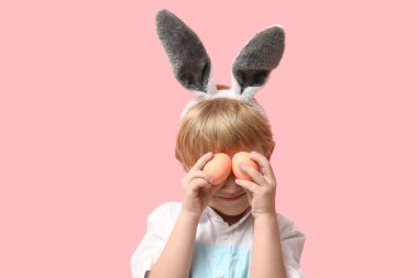 Pembe arka planda Paskalya yumurtaları olan tavşan kulaklı küçük bir çocuk.