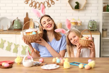 Sevimli küçük kız ve annesi tavşan kulaklı ellerinde sepetler dolusu Paskalya pastası ve mutfakta boyanmış yumurtalar tutuyorlar.
