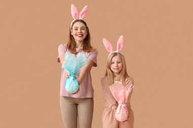 Şirin küçük kız ve annesi tavşan kulaklı ve kahverengi arka planda Paskalya hediyesi yumurtaları var.