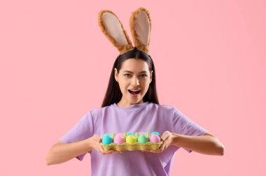 Pembe arka planda Paskalya yumurtaları olan tavşan kulaklı genç bir kadın.