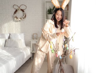 Genç Asyalı kadın yatak odasında söğüt dallarını paskalya yumurtalarıyla süslüyor.