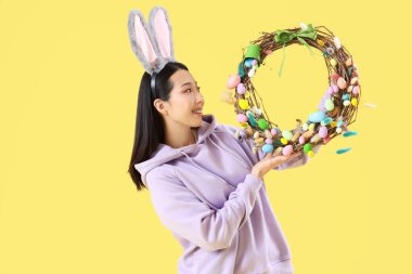 Sarı arka planda Paskalya çelengi olan tavşan kulaklı genç Asyalı kadın.
