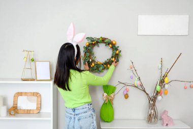 Tavşan kulaklı genç Asyalı kadın evdeki duvara Paskalya çelengi asıyor.