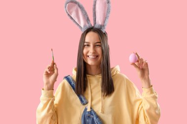 Tavşan kulaklı, boya fırçası ve pembe arka planda Paskalya yumurtası olan genç bir kadın.