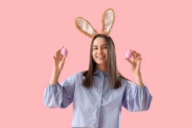 Pembe arka planda Paskalya yumurtaları olan tavşan kulaklı genç bir kadın.