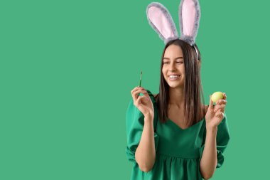 Boya fırçası ve yeşil arka planda Paskalya yumurtası olan tavşan kulaklı genç bir kadın.