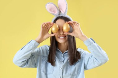 Sarı arka planda Paskalya yumurtaları olan tavşan kulaklı genç bir kadın.