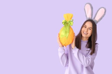 Leylak arka planında Paskalya hediyesi yumurtası olan tavşan kulaklı genç bir kadın.