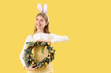 Sarı arka planda Paskalya çelengi olan tavşan kulaklı genç bir kadın.