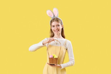 Sarı arka planda Paskalya yumurtası sepeti olan tavşan kulaklı genç bir kadın.