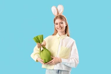 Mavi arka planda Paskalya hediyesi yumurtası olan tavşan kulaklı genç bir kadın.