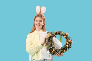 Mavi arka planda Paskalya çelengi olan tavşan kulaklı genç bir kadın.