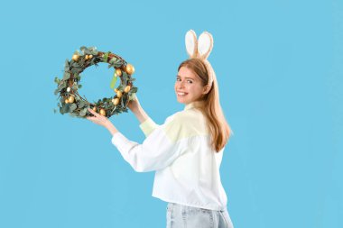 Mavi arka planda Paskalya çelengi olan tavşan kulaklı genç bir kadın.