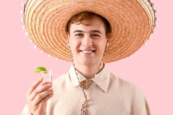 Portrait of young Mexican man in sombrero and with tequila on pink background