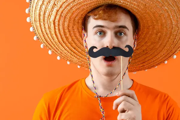 Sombrero giymiş, turuncu arka planda kağıt bıyıklı, şaşırmış Meksikalı bir adamın portresi.