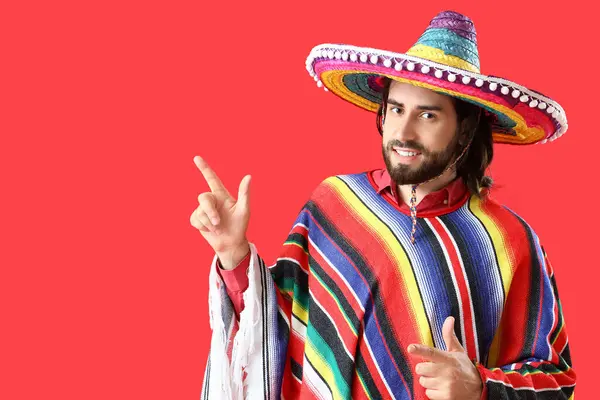 Handsome young Mexican man in sombrero and poncho pointing at something on red background