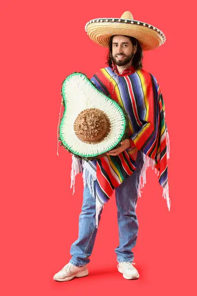 Kırmızı arka planda avokado şeklinde pinyatası olan yakışıklı Meksikalı genç adam.