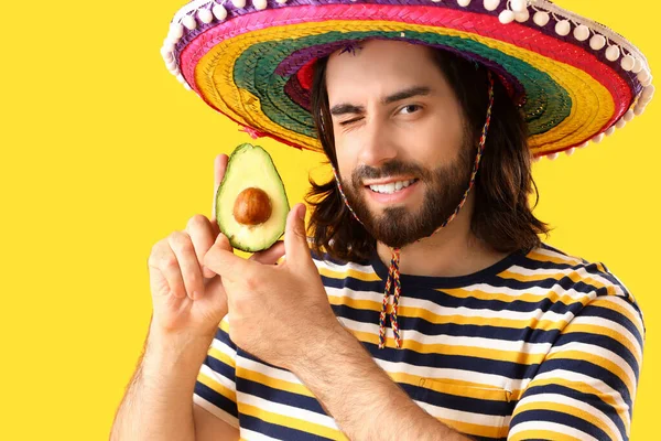 Handsome young Mexican man in sombrero and with avocado on yellow background