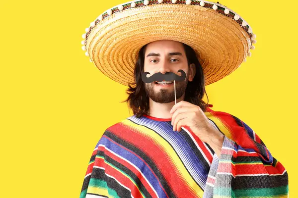 Sombrero giymiş, sarı arka planda kağıt bıyıklı genç bir Meksikalı.