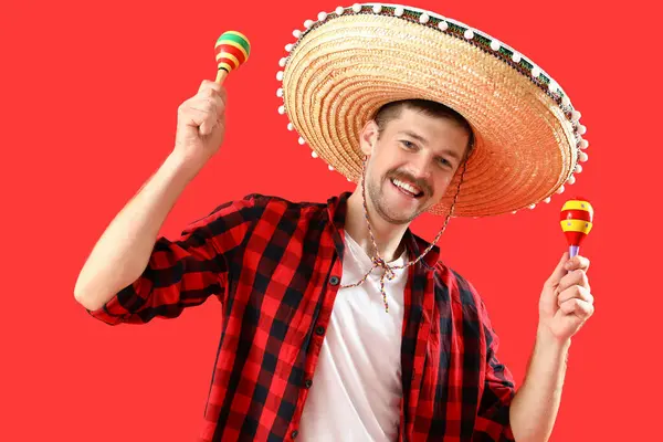 Happy young Mexican man in sombrero and with maracas on red background