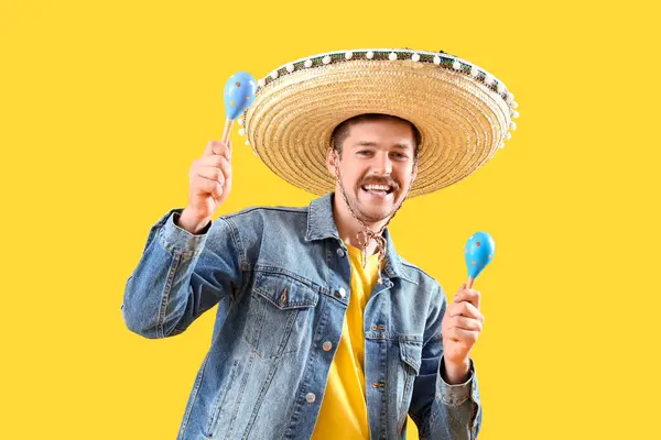 Happy young Mexican man in sombrero and with maracas on yellow background