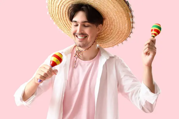 Young Mexican man in sombrero and with maracas on pink background