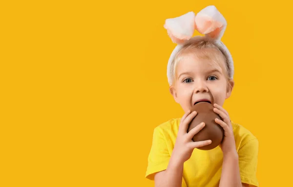 可爱的小女孩在兔子耳朵里吃黄色背景的巧克力蛋 复活节庆祝活动 — 图库照片