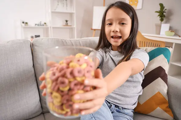 Cute little Asian girl with jar of cereal rings sitting on sofa at home