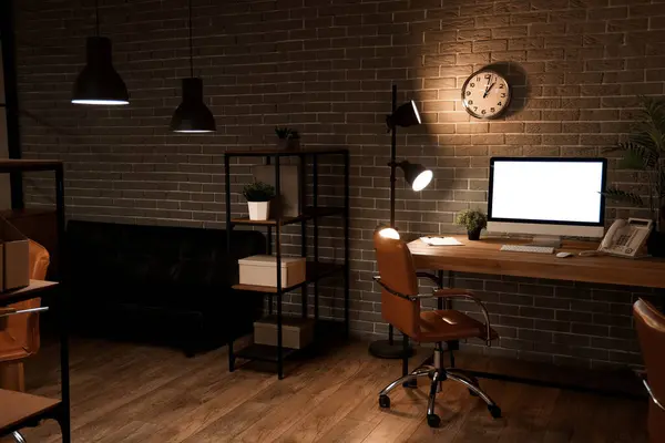 Interior of dark office with workplace, blank monitor and glowing lamps at night
