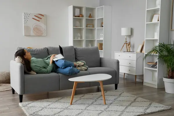 Genç bir kadın oturma odasındaki gri koltukta dizüstü bilgisayar kullanıyor.